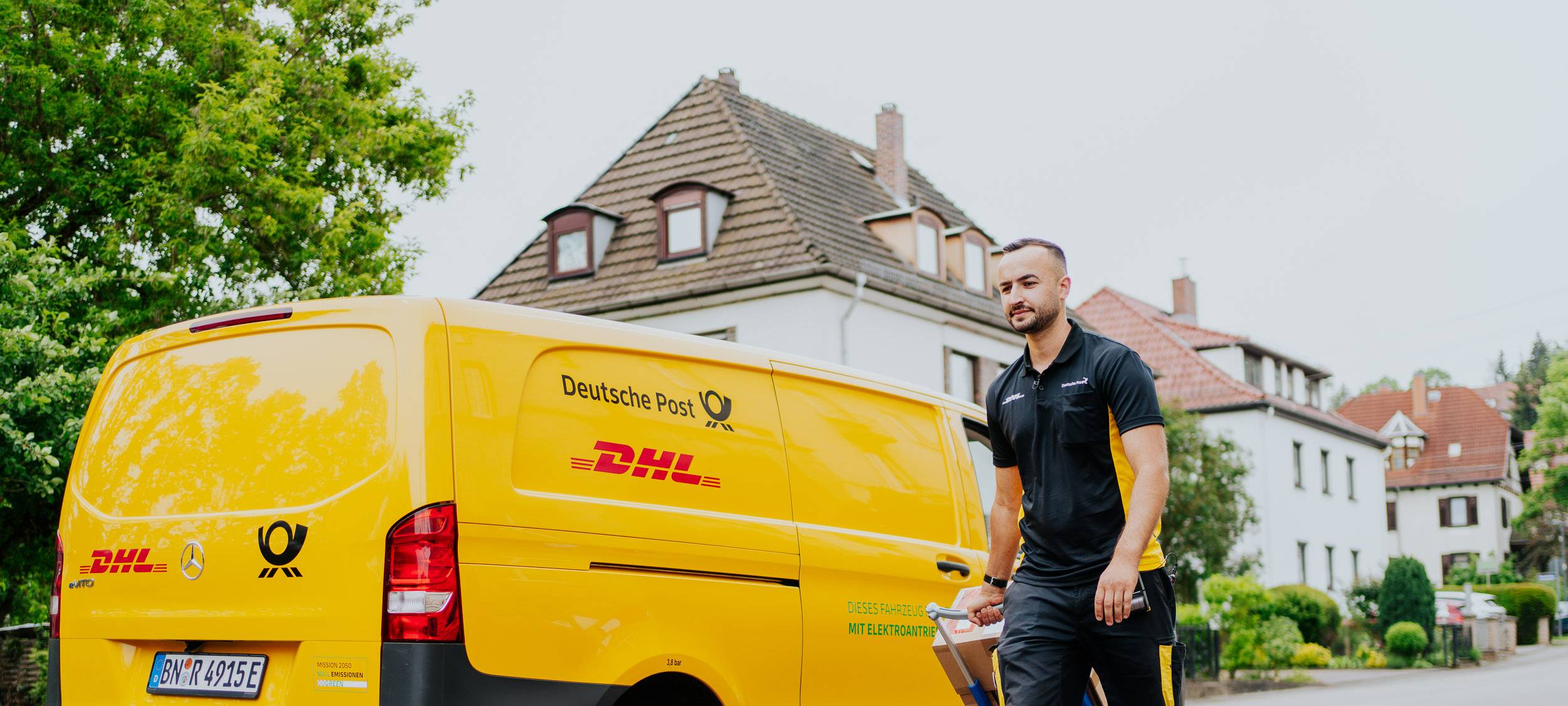 Ein männlicher Paketzusteller läuft neben dem gelben Auto der DHL entlang.